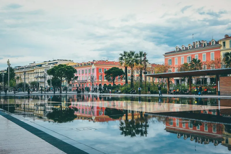 Καθρεφτισμένα νερά στο Promenade du Paillon στη Νίκαια, με φοινικόδεντρα, πολύχρωμα κτίρια και περαστικούς να χαλαρώνουν γύρω από το πάρκο σε σκηνικό που συνδυάζει αστική ζωντάνια και φυσική ομορφιά.